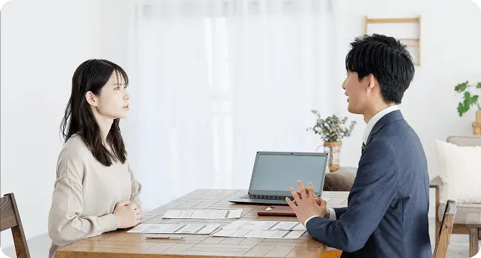 自宅のダイニングテーブルで、スーツ姿の男性が女性に資料を見せながら相談を行っている様子。